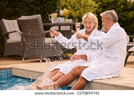 Senior Couple In Robes Outdoors Drinking Champagne By Swimming Pool On Splashing In Water On Spa Day
