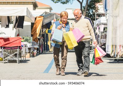 Senior Couple Shopping Together At Flea Market With Wife Watching In Husband Bags - Active Elderly Concept With Mature Man And Woman Having Fun In City - Happy Retired People Moments On Vivid Colors