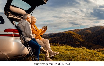 Senior couple sitting against the car, resting after hiking in countryside making selfie with smartphone. - Powered by Shutterstock