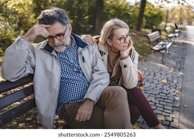 Senior couple sitting apart on a park bench in an autumn setting, looking upset and distant, portraying disagreement, relationship challenges, or emotional disconnect - Powered by Shutterstock