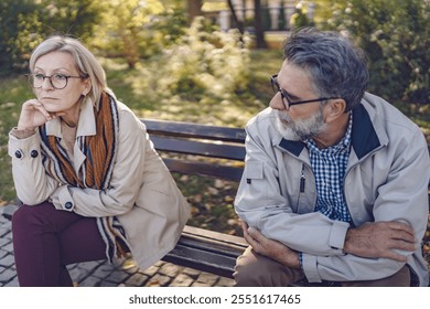 Senior couple sitting apart on a park bench in an autumn setting, looking upset and distant, portraying disagreement, relationship challenges, or emotional disconnect - Powered by Shutterstock