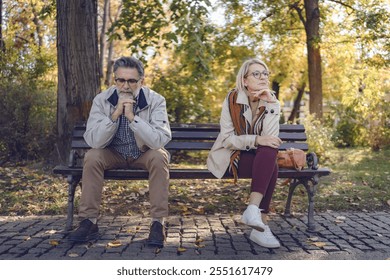 Senior couple sitting apart on a park bench in an autumn setting, looking upset and distant, portraying disagreement, relationship challenges, or emotional disconnect - Powered by Shutterstock