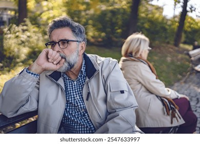 Senior couple sitting back-to-back on a park bench, looking upset and distant, depicting disagreement, relationship challenges, or emotional disconnect in an autumn park setting. - Powered by Shutterstock