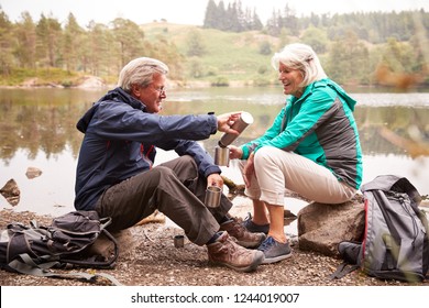 Senior Couple Sitting By A Lake Pouring Coffee From A Flask During A Camping Holiday, Lake District, UK