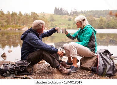 Senior Couple Sitting By A Lake Drinking Coffee During Camping Holiday Making A Toast With Their Mugs, Lake District, UK