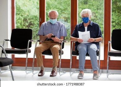 Senior couple sitting with face masks in a bright waiting room of  a hospital or an office - the man looks at a smartphone