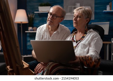 Senior Couple Sitting In Front Of Easel Holding Laptop Looking At Online Tutorials To Improve Drawing Technique. Retired Man And Woman Looking For Inspiration On Social Media Using Portable Computer.