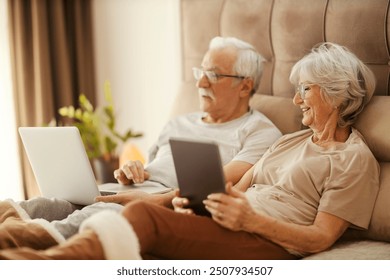 Senior couple sitting on bed in bedroom with tablet and laptop and using it for surfing online. - Powered by Shutterstock