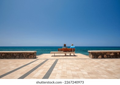 A senior couple sitting on a bench on the beach in the midday sun - Powered by Shutterstock
