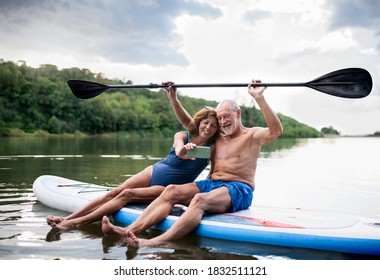 Senior couple sitting on paddleboard on lake in summer, taking selfie with smartphone.