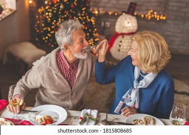 Senior Couple Sitting At A Table By A Nicely Decorated Christmas Tree, Having Christmas Dinner With Family, Trying Each Others Food