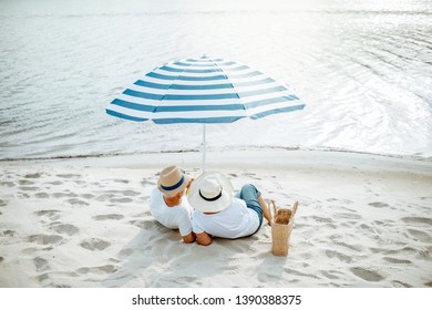 Senior Couple Sitting Together Under Umbrella On The Sandy Beach, Enjoying Their Retirement Near The Sea, Rear View