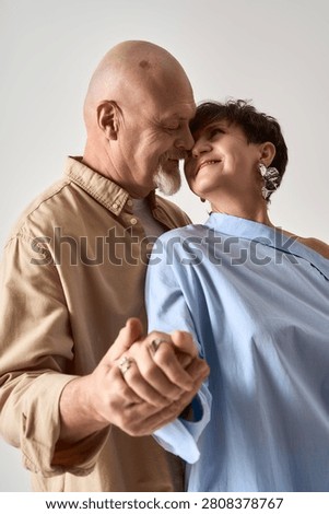 A senior couple smiles endearingly while holding hands, dressed fashionably and radiating love.