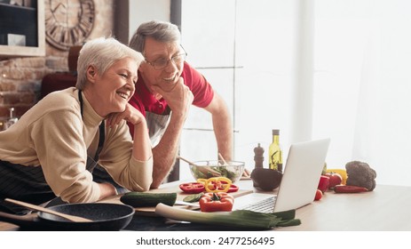 A senior couple smiles as they use a laptop to order groceries online while cooking in their kitchen. The woman holds a credit card in her hand, and they are surrounded by fresh produce. - Powered by Shutterstock