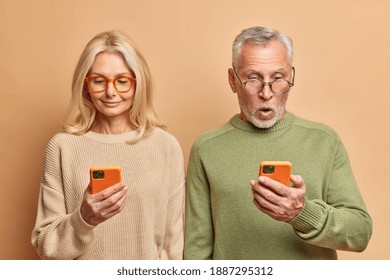 Senior Couple Stand Shoulder To Shoulder Use Smartphones For Browsing Internet Read Media Website Dressed In Casual Jumpers Isolated Over Brown Background. Substitute For Real Life. Tech Users