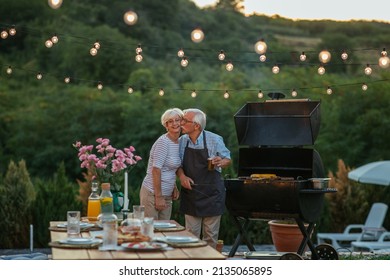 Senior couple standing in the backyard and preparing family gathering. Senior man kissing his wife who arranged table while he barbecuing