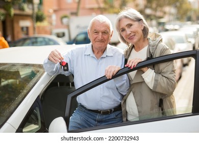 Senior Couple Standing Beside Their Car With Opened Door And Looking In Camera. Old Man Holding Keys In Hand.