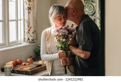 Senior couple standing in kitchen holding a bunch of flowers. Senior man kissing his wife holding her hand at home. - Powered by Shutterstock