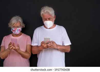 Senior Couple Standing On Black Background With Smartphone In Hand, Wearing Medical Mask Due To Coronavirus. The Phone Combines With The Color Of The Clothes. Older People Who Appreciate Modernity