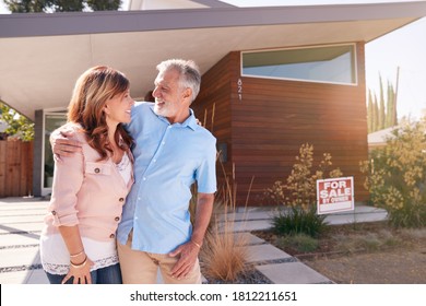 Senior Couple Standing Outdoors In Front Of House With For Sale Sign In Garden