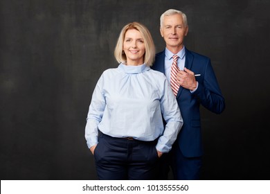 Senior Couple Standing Together At Dark Background. Beautiful Blond Woman Looking At Camera And Smiling While Elegant Old Man Standing Next To Her. 