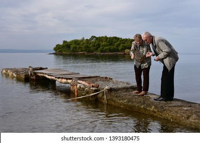 Senior Couple Stares At The Water Holding Hands On An Old Mooring Dock. Calm Nostalgic Sea. Small Islet And A Fishing Boat Seen At Distance.