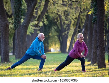 Senior Couple Stretching Legs And Warming Up For Exercise And Jogging In Park On Sunny Autumn Day