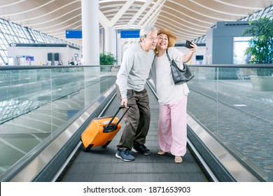 Senior couple taking a selfie photo together by using a smartphone while holding a luggage on the escalator of airport terminal - Powered by Shutterstock