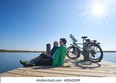 Senior couple with their bikes, Senior woman and man at rest on bike trip sitting on a boardwalk at lake                  - Powered by Shutterstock