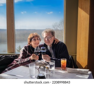 Senior Couple Toasting Each Other In Restaurant; Window With River View And Forest In Background