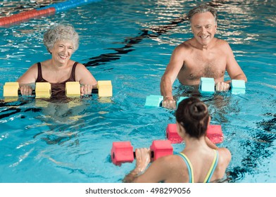 Senior couple in training session of aqua aerobics using dumbbells in swimming pool. Mature man and old woman practicing aqua fitness together. Healthy and fit senior couple doing aqua aerobics.