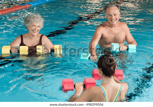 Senior couple in training session of aqua\
aerobics using dumbbells in swimming pool. Mature man and old woman\
practicing aqua fitness together. Healthy and fit senior couple\
doing aqua aerobics.