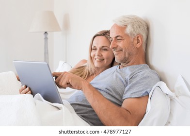 Senior couple using digital tablet while lying in bed at home. Happy senior couple surfing on the tablet in bedroom. Retired man and smiling woman planning on laptop their summer vacation. - Powered by Shutterstock