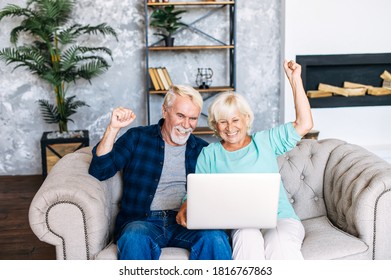 Senior Couple Is Using Laptop For Watching Sports Match Or Online Lottery Together Sitting On Comfortable Sofa In Cozy Living Room. Cheerful Older Spouses Holding Hands Up In Victory Gesture, Triumph