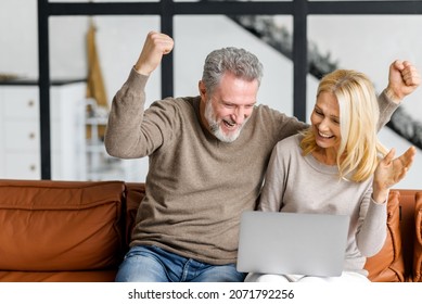 Senior Couple Is Using Laptop For Watching Sports Match Or Online Lottery Together Sitting On Comfortable Sofa In Living Room. Cheerful Middle-aged Spouses Raising Hands Up In Victory Gesture, Triumph