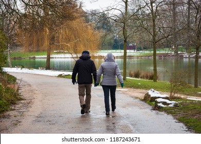 Senior Couple Walking In Park Near Daumesnil Lake (Paris, France) On Wet Road In Rare Snowy Day. 