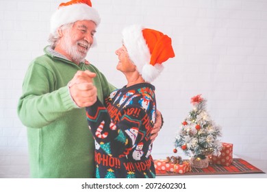 Senior Couple In Warm Clothing And Santa Hat Holding Each Others Hands And Dancing In Front Of Decorated Christmas Tree At Home. Loving Old Romantic Heterosexual Couple Celebrating Christmas Together