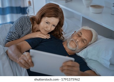 Senior Couple Watching Video With Digital Tablet In The Dark Before Sleeping. Mature Man Showing Digital Tablet To Wife While Lying On Bed Together Late In Night. Sleepy Couple Using Computer.