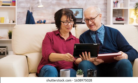 Senior Couple Waving At Tablet During A Video Call. Elderly People Sitting On Sofa.