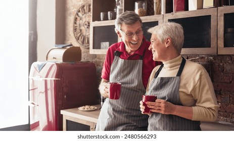 A senior couple wearing aprons stands in their kitchen, smiling and enjoying their coffee together. The man has a red shirt on and glasses. The woman has on a cream sweater, copy space - Powered by Shutterstock