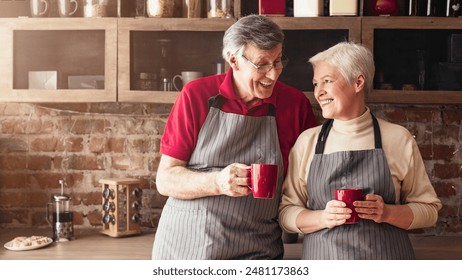 A senior couple wearing aprons stands together in a kitchen, each holding a red mug of coffee. They are smiling and looking at each other, enjoying a moment of connection and togetherness. - Powered by Shutterstock