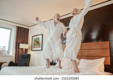 Senior couple in white robes jumping on bed and looking happy - Powered by Shutterstock