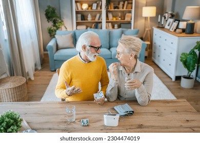 Senior couple woman take medicine or supplement vitamin treatment cure while her husband old caucasian man sit beside at the table at home real people family old people retirement and health concept - Powered by Shutterstock