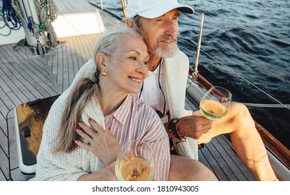 Senior Couple Wrapped With Plaid And Sitting On A Yacht Deck With Bocals Of Wine. Two Loving Mature People Sitting Together On A Sailboat And Looking Away.