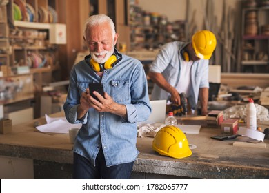 Senior craftsman is taking a break from work. He is using his mobile phone and smiling. His apprentice is drilling wood in the background. - Powered by Shutterstock