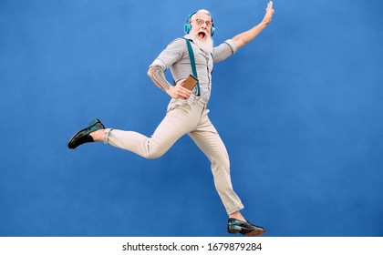 Senior Crazy Man Jumping While Listening Music With Wireless Headphones - Hipster Old Guy Having Fun Dancing Outdoor - People Happiness And Elderly Technology Addicted Concept - Blue Background