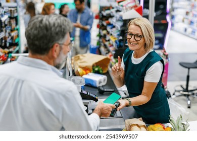 Senior customer using smartphone to make contactless payment at grocery store checkout, assisted by friendly cashier pointing to pos terminal - Powered by Shutterstock