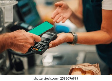 Senior customer using smartphone to make contactless payment at grocery store checkout, holding phone near payment terminal while cashier holds terminal and assists with transaction