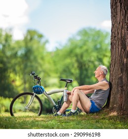 Senior cyclist sitting by a tree in a park with a water bottle in his hand and listening to music on headphones shot with tilt and shift lens