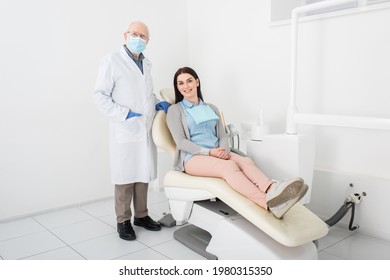 Senior Dentist In Medical Mask And Latex Gloves Standing Near Pretty Woman Sitting In Dental Chair In Clinic